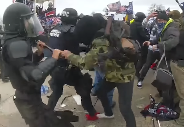 Police body camera footage from January 6th showing a chaotic close-quarters struggle between a Metropolitan Police officer in riot gear and a rioter in camouflage attempting to seize a baton. In the background, a large crowd with flags clashes with a police line.