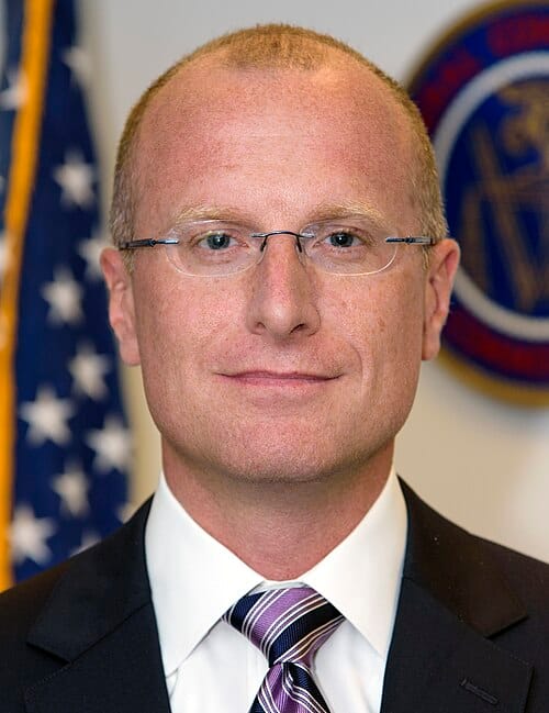 Official government headshot of FCC Chairman Brendan Carr; he wears rimless glasses and a sharp dark suit.